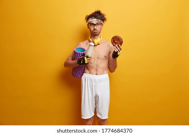 Indoor Shot Of Displeased Sportsman Feels Temptation To Eat Junk Food, Carries Foam Roller, Wants To Have Perfect Body, Wears Stereo Headphones Around Neck Shorts, Headband, Sport Gloves, Does Workout