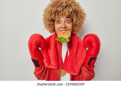 Indoor Shot Of Female Boxer Eats Appetizing Burger After Fight Feels Very Hungry Won Golden Medal On Comptetion Dressed In Uniform Stares At Camera Isolated Over Grey Background. Sport And Fast Food