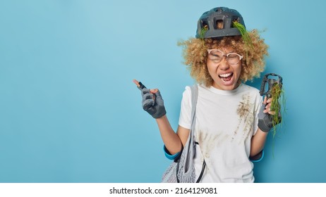 Indoor shot of female cyclist fell off bike while riding wears safety helmet elbow pads gloves screams loudly wears white t shirt smeared with dirt isolated over blue background points at empty space