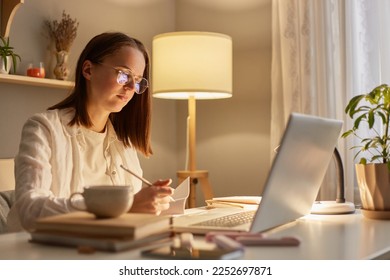 Indoor shot of focused woman wearing white shirt sitting on table and working on laptop near window, having concentrated facial expression, studying hard or works with a new project. - Shutterstock ID 2252697871