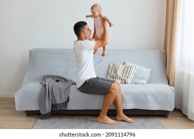 Indoor Shot Of Ghandsome Brunette Father Holding Infant Baby Girl In Hands And Understand They Need Changing Baby's Diaper, Feeling Bad Smell, Raising Kid Up Air.