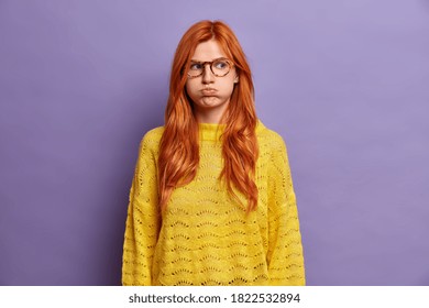 Indoor shot of ginger offended young woman blows cheeks and makes unhappy grimace holds air has moody expression wears yellow sweater displeased with awful situation happened shows bad character