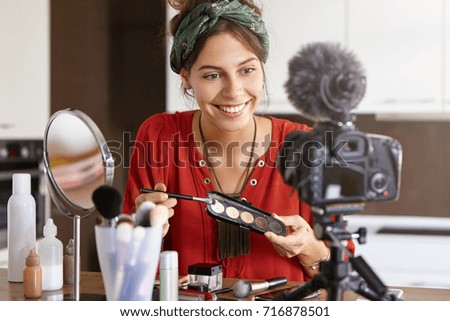 Indoor shot of glad woman sits at home, uses cosmetic products for making video diary, looks at camera with appealing smile. Elegant female applies eyeshadow, shows for women its great effect