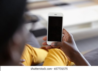 Indoor shot of hand of dark-skinned man holding cell phone with blank copy space screen for your text or advertising content while lying at couch at home searching internet. People and technology