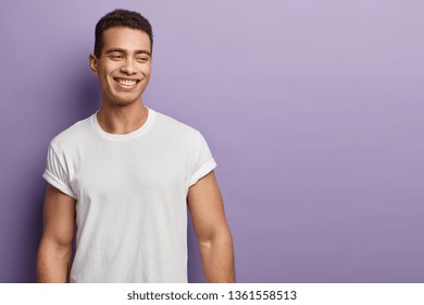 Indoor Shot Of Handsome Muscular Guy With Positive Expression, Wears Casual Mock Up White T Shirt For Your Promotion, Stands Against Purple Background, Free Space Aside. People And Emotions.