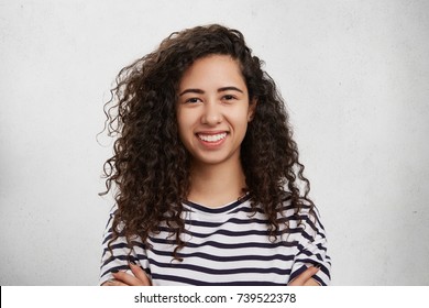 Indoor shot of happy beautiful woman with curly bushy dark hair, healthy pure skin, broad smile and white even teeth, looks directly into camera, glad to hear positive news. Positiveness concept