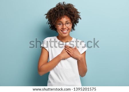 Indoor shot of happy dark skinned lady swears or promises something, holds hands on chest, tells truth, being honest, looks at camera friendly, has charming smile, wears casual white t shirt, glasses