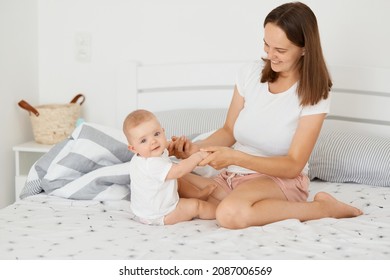 Indoor Shot Of Happy Family Posing In Bedroom At Home, Infant Baby Sitting Near Mother And Looking At Camera, Young Adult Woman Holding Baby's Hands, Expressing Love.