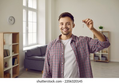 Indoor Shot Of Happy Home Owner With Key To His New House. Cheerful Handsome Single Young Caucasian Man In Casual Shirt Standing In Living Room, Showing Keys And Smiling. Real Estate, Purchase Concept