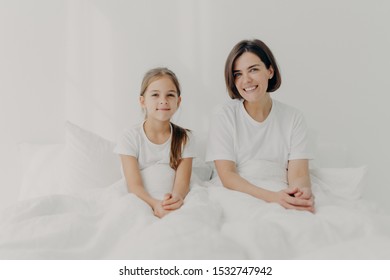 Indoor Shot Of Happy Mom And Small Daughter Feel Relaxed And Pleased Pose In Bed, Enjoy Comfort, Spend Leisure Time At Home, Wake Up Late, Have Enough Sleep, Dressed Casually. Morning Concept