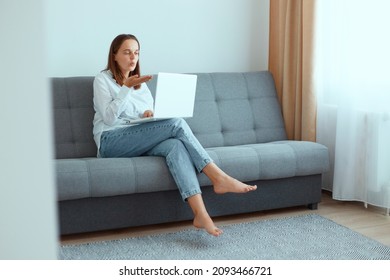 Indoor Shot Of Happy Romantic Dark Haired Woman Sitting On Sofa With Laptop, Wearing White Shirt And Jeans, Looking At Notebook Screen While Having Video Call With Husband, Sending Air Kissing.