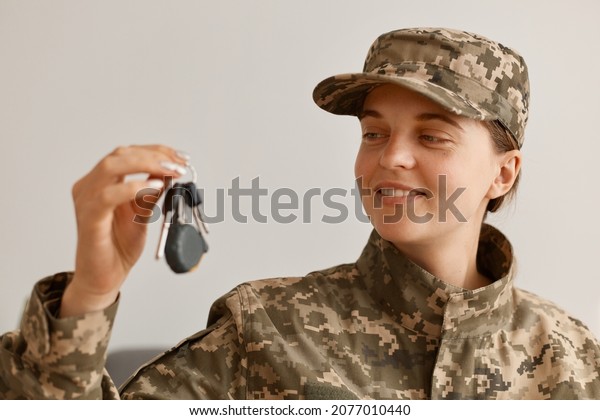 indoor shot of happy smiling woman\
soldier wearing camouflage uniform and hat, posing at home, holding\
and looking at key in hands, buying new flat or\
car.