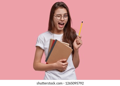 Indoor Shot Of Happy Young Caucasian Female Genius Gets Good Idea, Blinks Eye, Holds Pencil, Carries Notebooks, Has Fun Indoor, Dressed In White Casual T Shirt, Isolated Over Pink Background