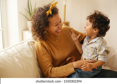 Indoor Shot Of Happy Young Hispanic Woman With Brown Wavy Hair Relaxing At Home Embracing Her Adorable Toddler Son. Cheerful Mother Bonding With Infant Son, Sitting On Sofa In Living Room, Laughing