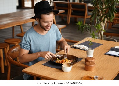 Indoor Shot Of Happy Young Student Wearing Stylish Hat Eating Delicious Food With Knife And Fork During Break At University Canteen, Enjoying Fresh And Healthy Lunch, Sitting At Wooden Table Alone