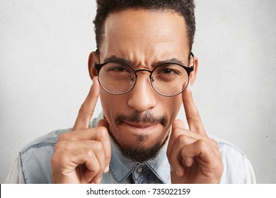 Indoor shot of hipster guy in round spectacles keeps fingers on temples, tries to concentrate and find right solution or way out in difficult situation, isolated over white studio background
