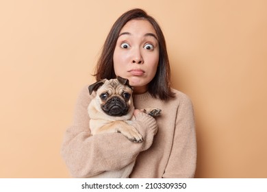 Indoor Shot Of Impressed Young Asian Woman Poses With Pug Dog Shocked To Finds Out Something Embarrassing Sees Something Breathtaking Poses Against Brown Background. Domestic Animals Concept