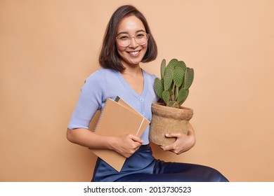 Indoor shot of intelligent Asian student holds potted cactus and notepads prepares for exam wears big spectacles casual jumper and skirt isolated over beige background looks gladfully at camera
