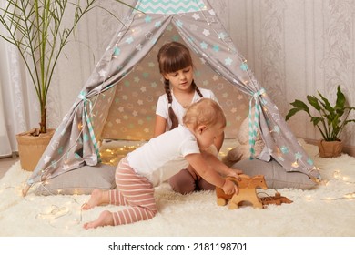 Indoor Shot Of Little Sisters Playing In Peetee Tent With Eco Soft Toy, Preschooler Girl And Toddler Baby Sitting On Floor Near Wigwam, Having Fun With Interest.