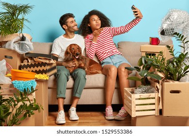 Indoor Shot Of Lovelly Family Couple Make Selfie Portrait, Afro Woman Blows Air Kiss In Camera Of Smartphone, Pose On Comfortable Sofa With Pet, Relocate In New Modern Apartment, Unpack Boxes Around