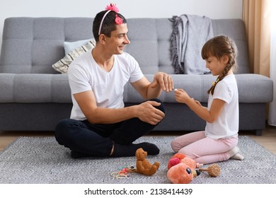 Indoor Shot Of Man Wearing White T Shirt Sitting On Floor And Playing With His Daughter, Father In Pink Hair Band With Flower Giving Hand To Kid For Painting Nails.