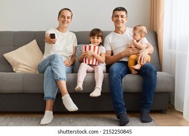 Indoor Shot Of Man, Woman And Their Daughter Posing At Home In Living Room And Watching Tv Together, Mother Holding Remote Control, Child With Popcorn, Enjoying Time Together.