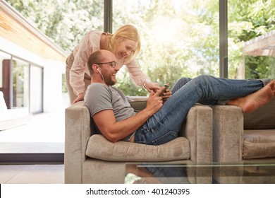 Indoor Shot Of Mature Couple At Home Using Digital Tablet And Smiling. Man Is Sitting On Sofa With Woman Standing By. Both Looking At Tablet Computer And Laughing.