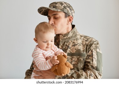 Indoor shot military man wearing camouflage uniform and cap, standing with his little infant daughter in hands, father missing his child and kissing baby, expressing love. - Powered by Shutterstock