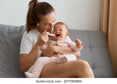 Indoor shot of mother measuring temperature of her sick crying baby sitting on sofa in living room, little infant child catching cold, having influenza or grippe.