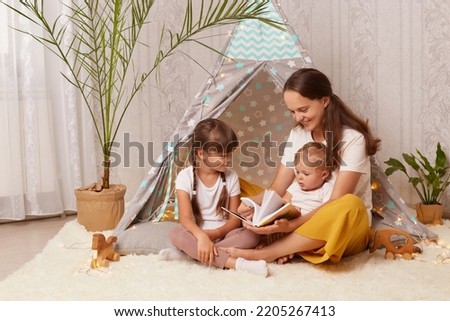 Indoor shot of mother with two charming little girls sitting together in wigwam, mom with her children reading interesting book with fairy tale in peetee tent.