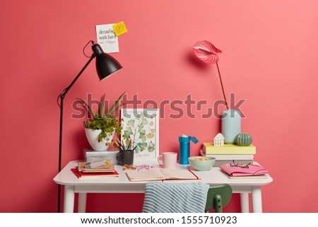 Indoor shot with no people, white desk of student with lamp, opened notebook, books, thermos of coffee, rosy calla lilies in vase pink wall sticky notes insctibed motivation phrases. Cozy workplace