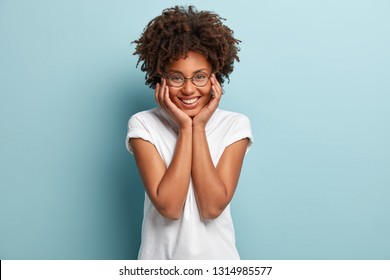 Indoor shot of peaceful tender woman touches cheeks with both hands, has broad smile, shows perfect teeth, wears spectacles and casual t shirt, isolated over light blue background. People and optimism - Shutterstock ID 1314985577