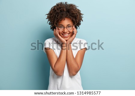 Indoor shot of peaceful tender woman touches cheeks with both hands, has broad smile, shows perfect teeth, wears spectacles and casual t shirt, isolated over light blue background. People and optimism