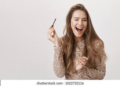 Indoor shot of positive attractive caucasian woman winking and smiling broadly while holding mascara and applying it, standing in evening dress over gray background. Girl gets ready to nightclub