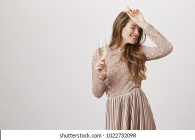 Indoor Shot Of Positive Attractive Young Female In Evening Dress With Glass Of Champagne, Holding Hand On Forehead And Smiling, Standing With Closed Eyes Over Gray Background. Girl Dances Sensually