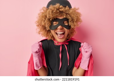 Indoor shot of positive curly haired woman clenches fists celebrates specialoccasion exclaims like winner wears protective helmet eyemask and superhero costume isolated over pink background. - Powered by Shutterstock