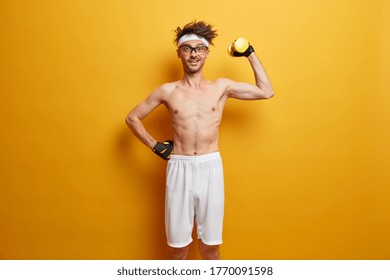 Indoor Shot Of Positive Man Holds Heavy Sport Equipment, Enjoys Workout In Own Gym, Has Skinny Body, Wants To Be Healthy And Fit, Wears Transparent Glasses, Sport Gloves, Poses Over Yellow Wall