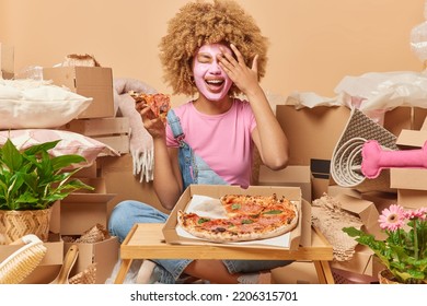 Indoor Shot Of Positive Woman Wears Beauty Facial Mask Covers Face With Palm Eats Delicious Pizza Has Fun Celebrates Moving Day Packs Personal Belongings In Cardboard Boxes. Relocation Concept