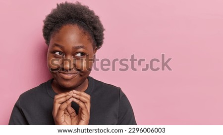 Indoor shot of pretty dark skinned woman schemes future plans steepls fingers has intention to do something looks with cunny expression aside wears casual black t shirt isolated on pink background