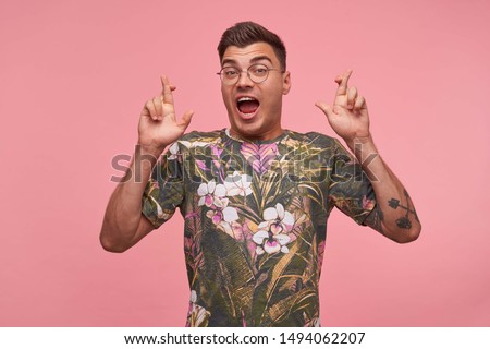 Indoor shot of pretty young guy raising hands with crossed fingers for good luck, being excited about something, looking to camera with raised eyebrowns and wide opened mouth