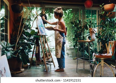 Indoor shot of professional female artist painting on canvas in studio with plants.
