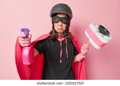 Indoor Shot Of Puzzled Displeased Female Superhero Holds Cleaning Detergent And Plunger Ready For Washing Dressed In Costume With Cloak Does Household Duties Isolated Over Pink Studio Background