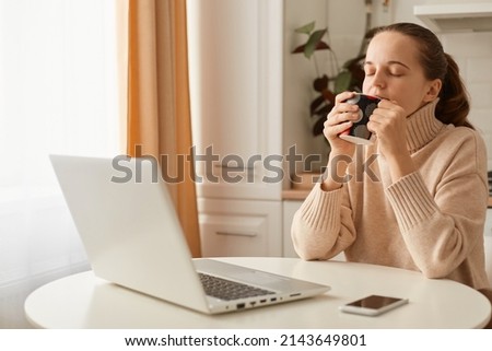 Indoor shot of relaxed woman wearing beige jumper posing in kitchen and working on laptop, having little rest and drinking and enjoying hot coffee or tea.
