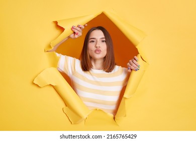 Indoor shot of romantic lovely woman wearing striped shirt sending air kissing to camera, expressing love and gentle, looking through hole in yellow paper.