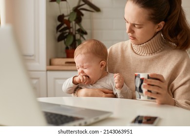 Indoor Shot Of Sad Dark Haired Woman Wearing Beige Sweater Posing In Kitchen With Her Crying Baby And Holding Cup Of Tea Or Coffee, Trying To Calm Down Kid.