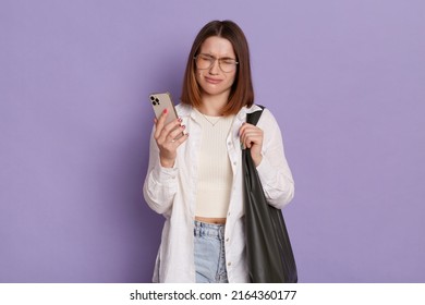 Indoor shot of sad depressed woman with black bag wearing white shirt and jeans posing isolated over purple background, standing with cell phone in hands and crying, having bad news.