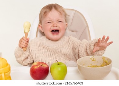 Captura de pantalla cubierta de triste bebé disgustada vestida con un saltador beige sentado en una silla y llorando, no quiere comer avena, aislada sobre fondo blanco