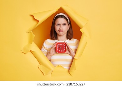 Indoor Shot Of Sad Upset Woman Wearing Hair Band And Striped T Shirt Standing In Yellow Paper Hole, Holding Red Alarm Clock, Looking At Camera Expressing Sorrow And Sadness.