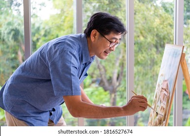 Indoor Shot Of Senior Asian Man Sit And Painting Picture On Easel Inside Of The House. Leisure Activity For Elderly For Relaxation,  Happy Retirement Lifestyle Concept.