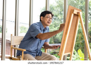 Indoor shot of senior Asian men using brush and oil color to paint on painting canvas. With smile on his face. Happy retirement concept for active senior. With Blurred living room background.